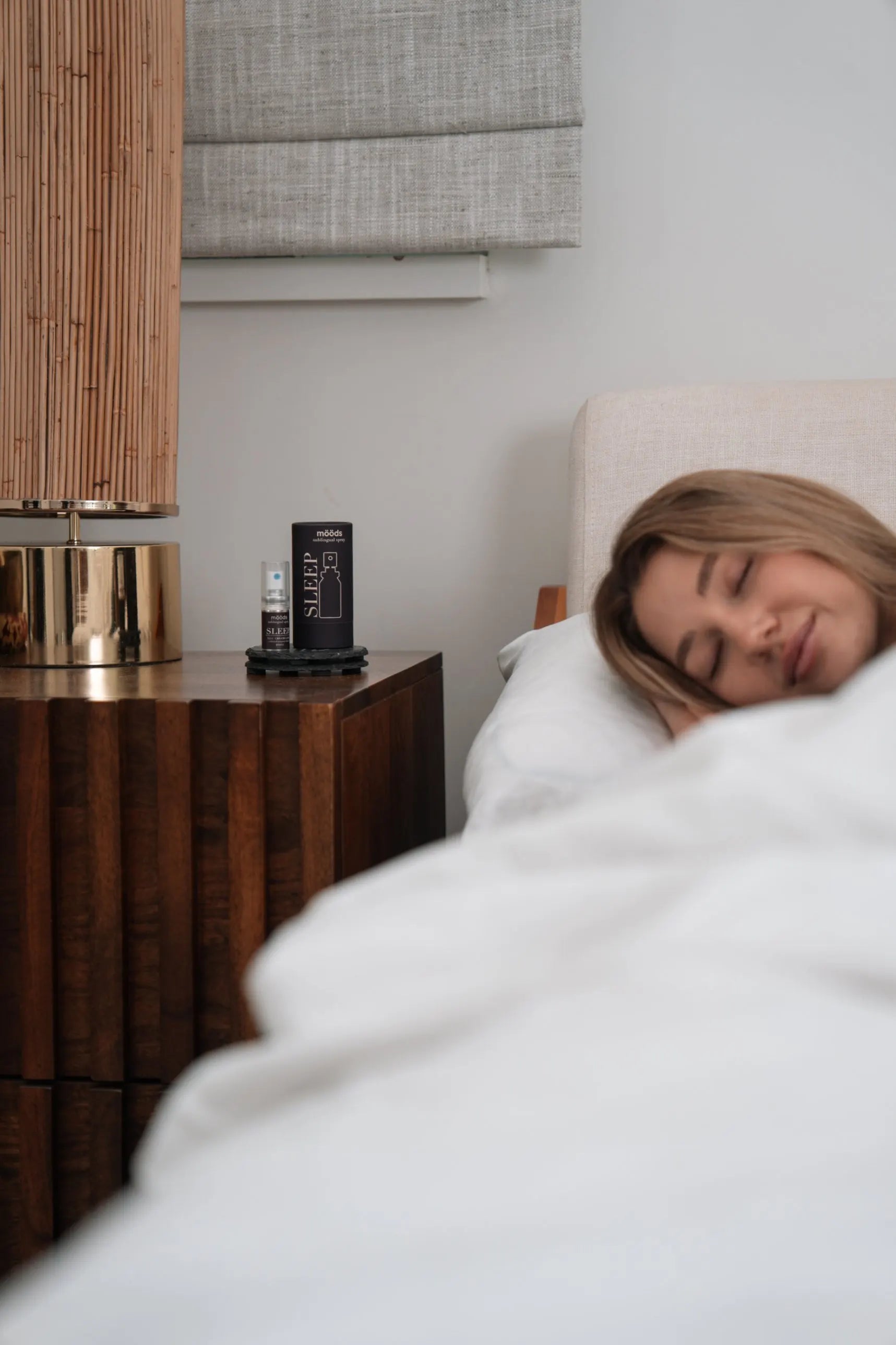 A woman asleep in bed; on the nightstand beside her, the SLEEP spray stands next to its outer tube